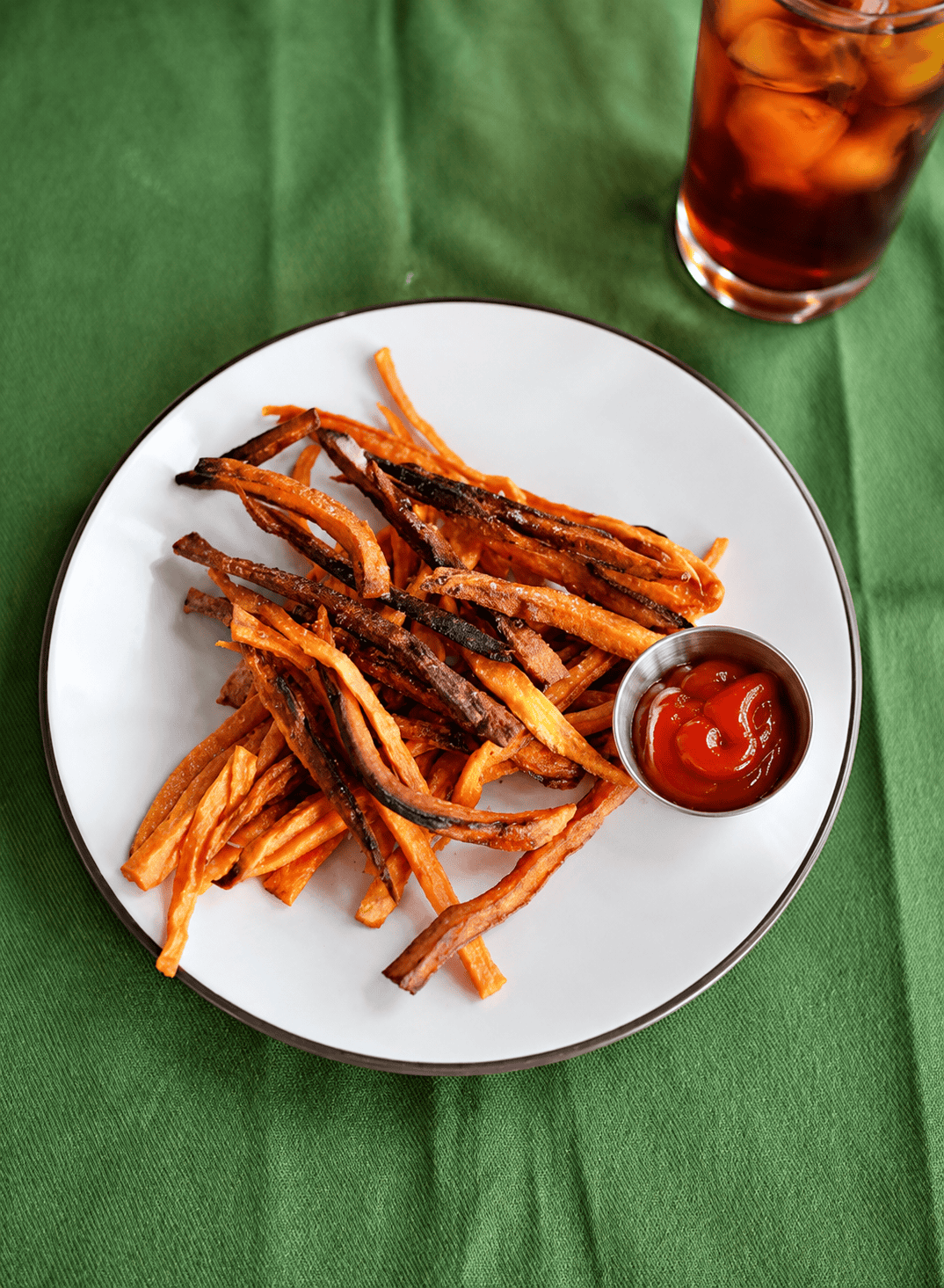 A plate of sweet potato fries with a mini cup of ketchup and a glass of iced tea.