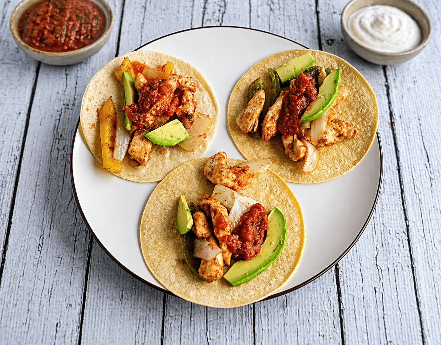 A plate with 3 fajitas on it with a bowl of sour cream and salsa behind the plate.
