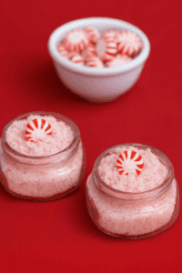 Two jars of pink sugar scrub with a bowl of peppermint candies behind the jars.