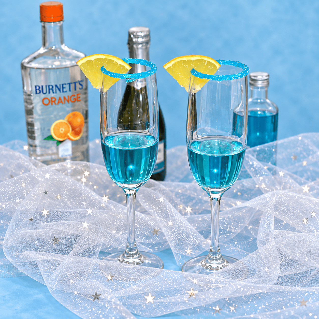 A bottle of vodka, a bottle of champagn, a bottle of blue curacuo, and two champagne glasses.