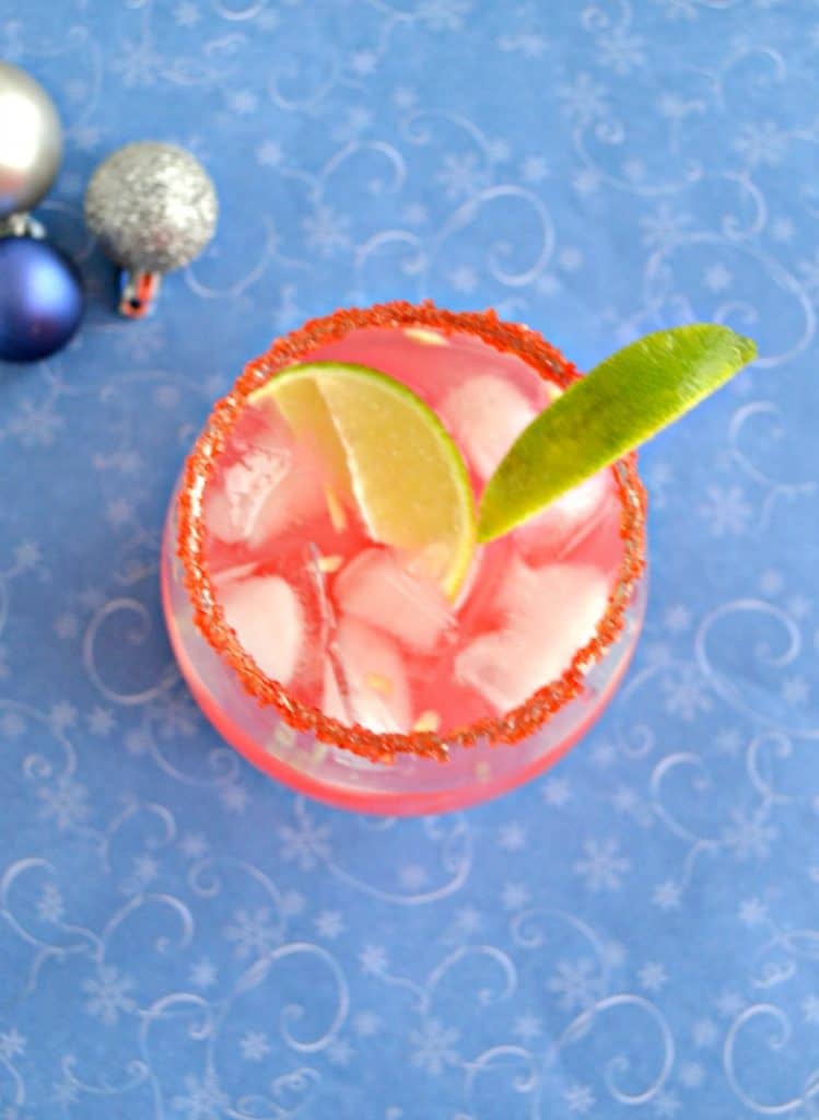 A top view of a glass filled with ice and pink liquid with red sugar around the rim and a lime wedge on a blue background.