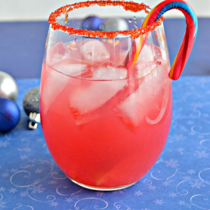 A stemless wineglass rimmed with red sugar, filled with pink liquid, with a candy cane swizzle stick, on a blue background.