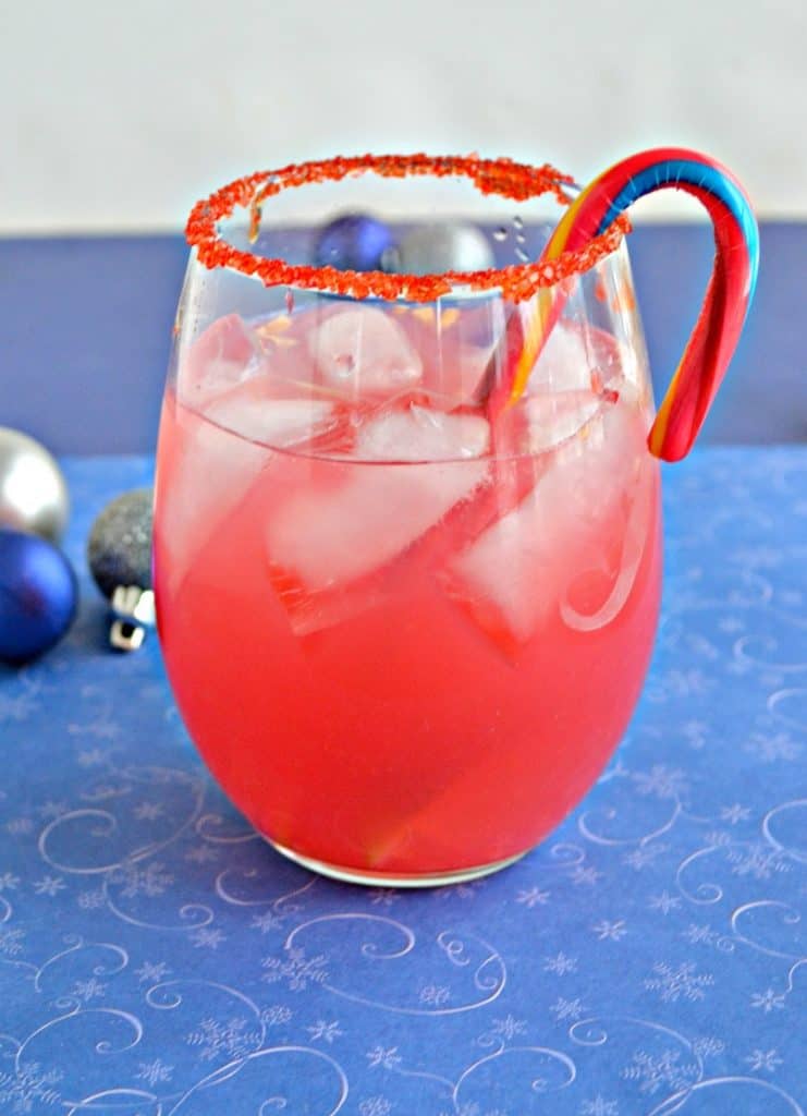 A stemless wineglass rimmed with red sugar, filled with pink liquid, with a candy cane swizzle stick, on a blue background.
