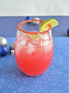 A stemless glass rimmed with red sugar with pink liquid inside and a lime wedge on the rim on a blue background.