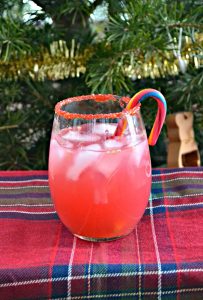 A stemless wineglass rimmed with red sugar, filled with pink liquid, with a candy cane swizzle stick, on a red plaid tablecloth with a Christmas tree behind it.