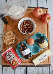 A cutting board with apples, a bowl of pecans, a cup of flour, a cup of brown sugar, and a stick of butter.