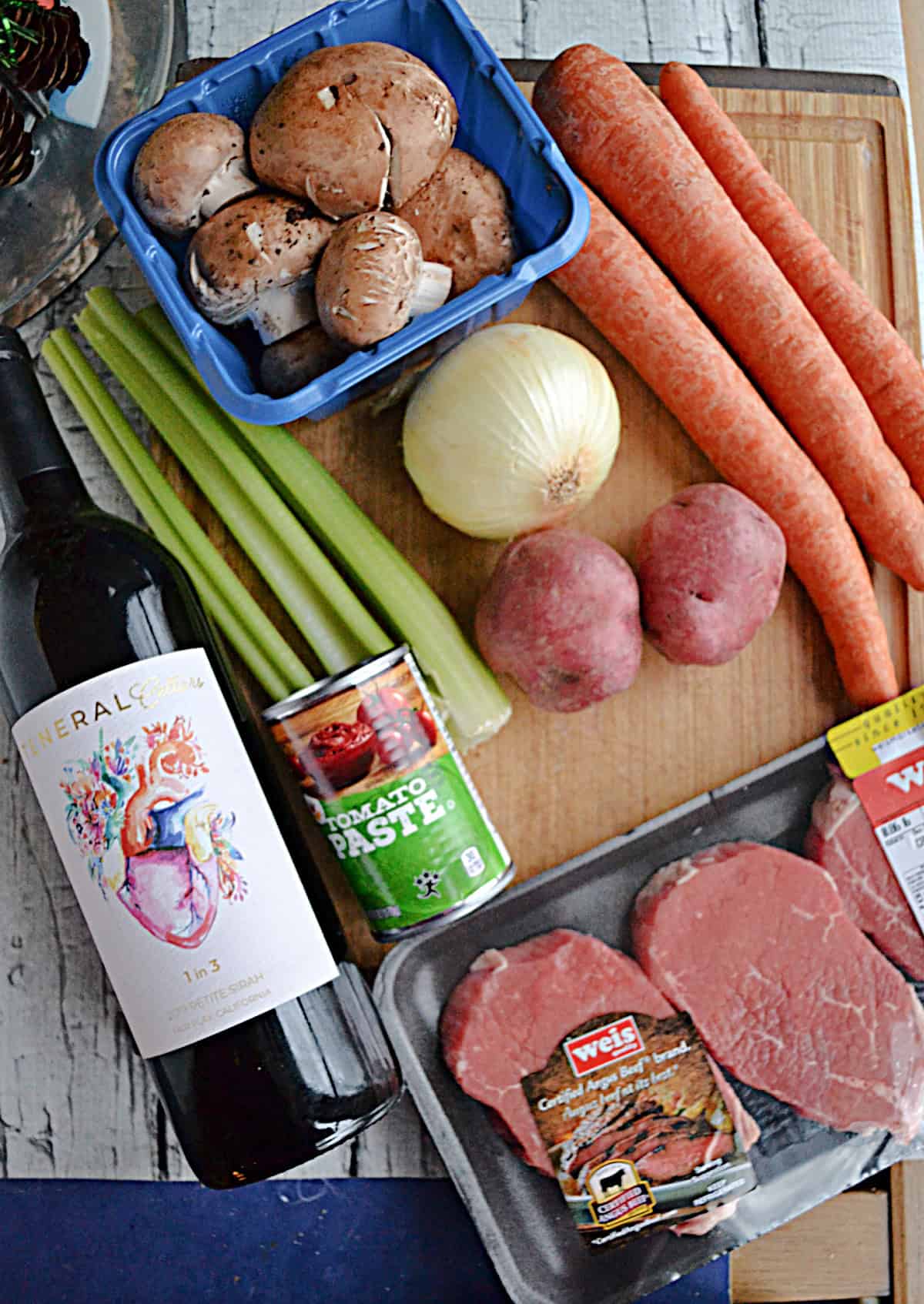 A cutting board with a bottle of wine, beef steaks, carrots, onions, celery, and mushrooms on it.