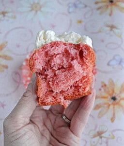A hand holding a pink velvet cupcake with a bite taken out of it.