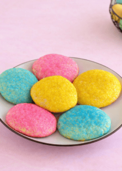A plate of pastel colored cookies with a basket of Easter eggs in the background.