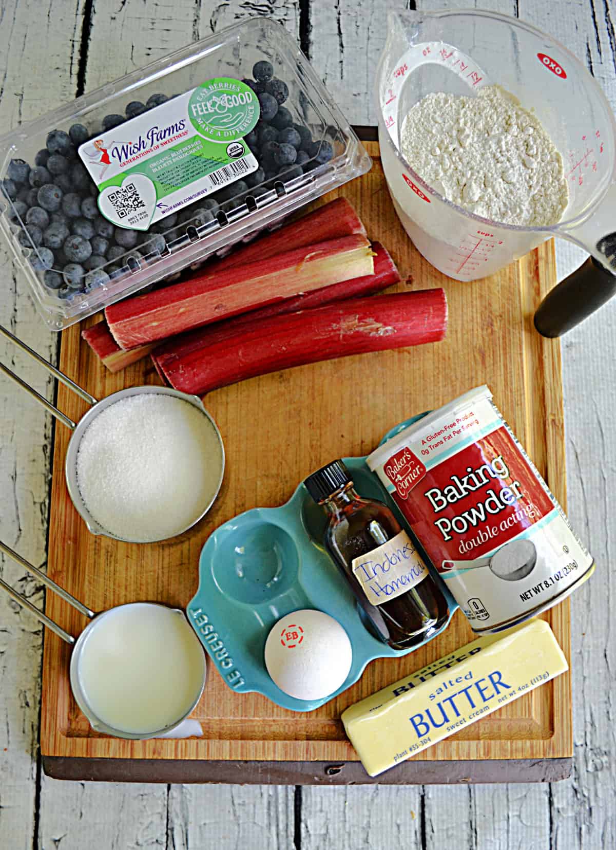 A cutting board with a container of blueberries, some rhubarb, flour, sugar, butter, eggs, and vanilla.
