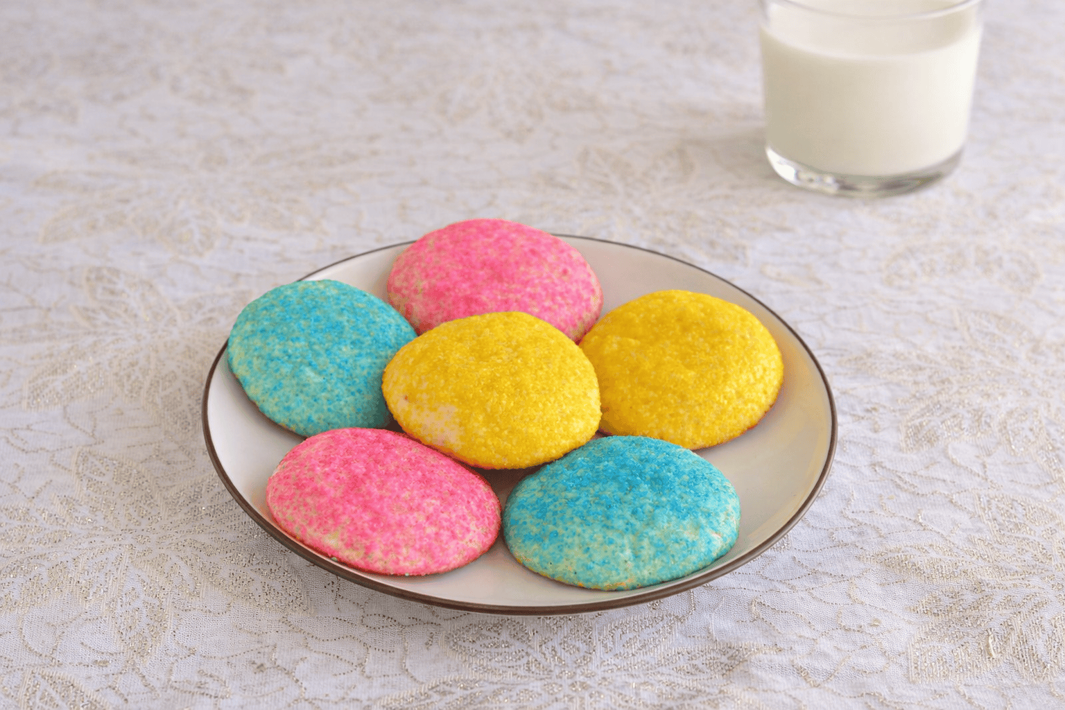 A plate of pastel colored cookies with a glass of milk behind the plate.