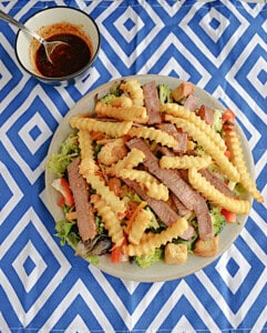 A salad topped with steak and fries with a bowl of dressing behind it.