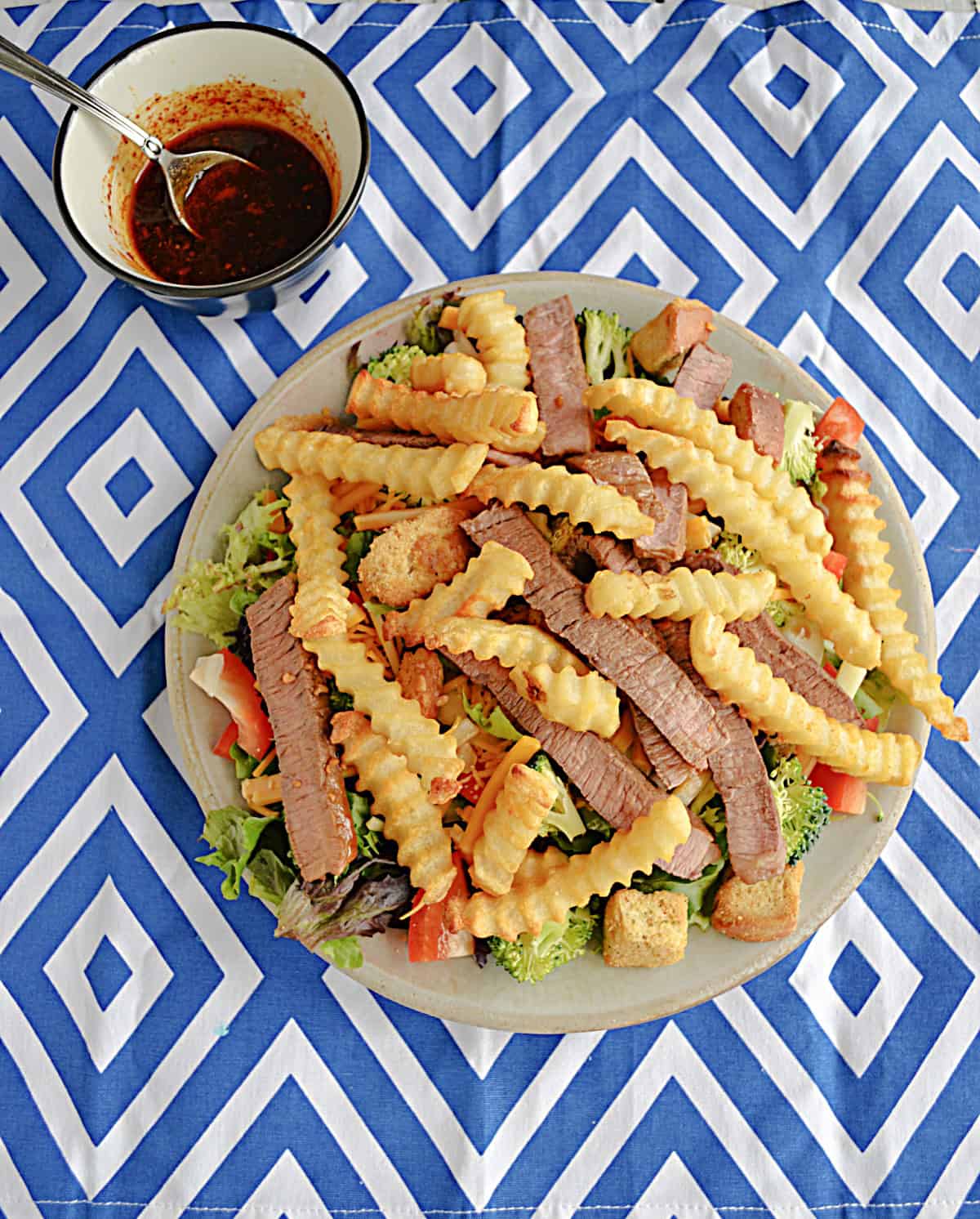 A salad topped with steak and fries with a bowl of dressing behind it.