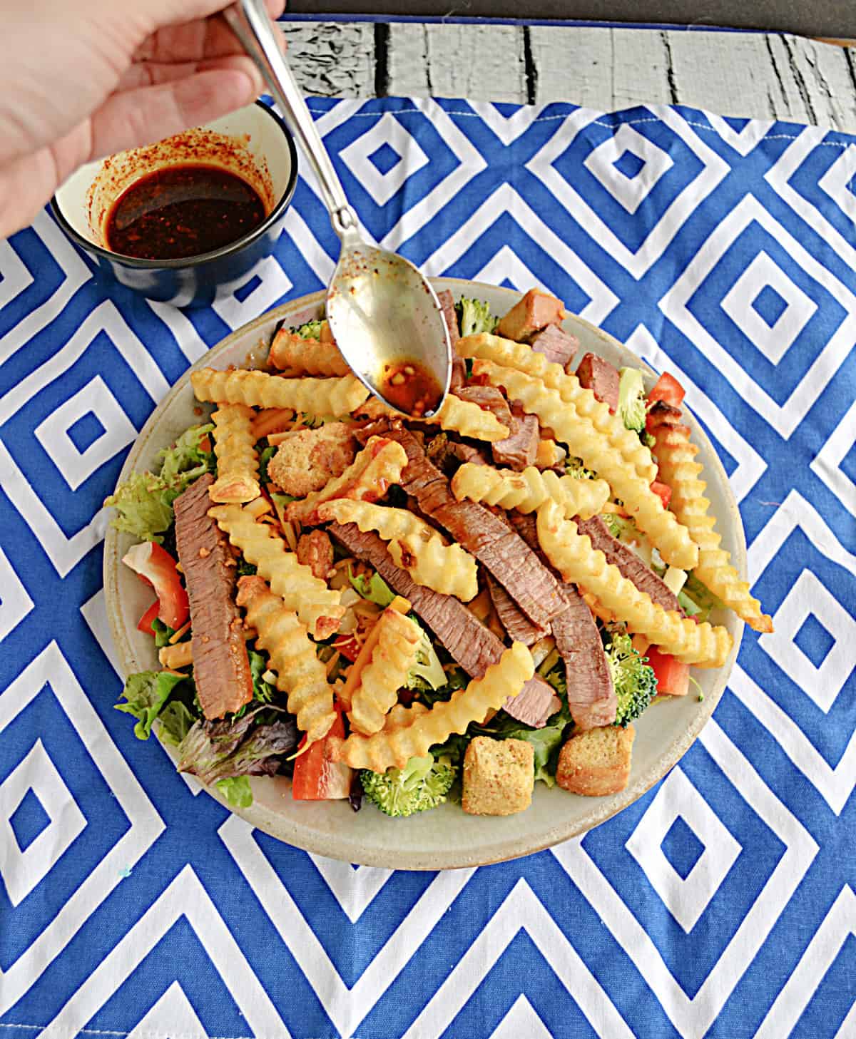 A salad topped with fries and steak and a spoon drizzling dressing over top of it.