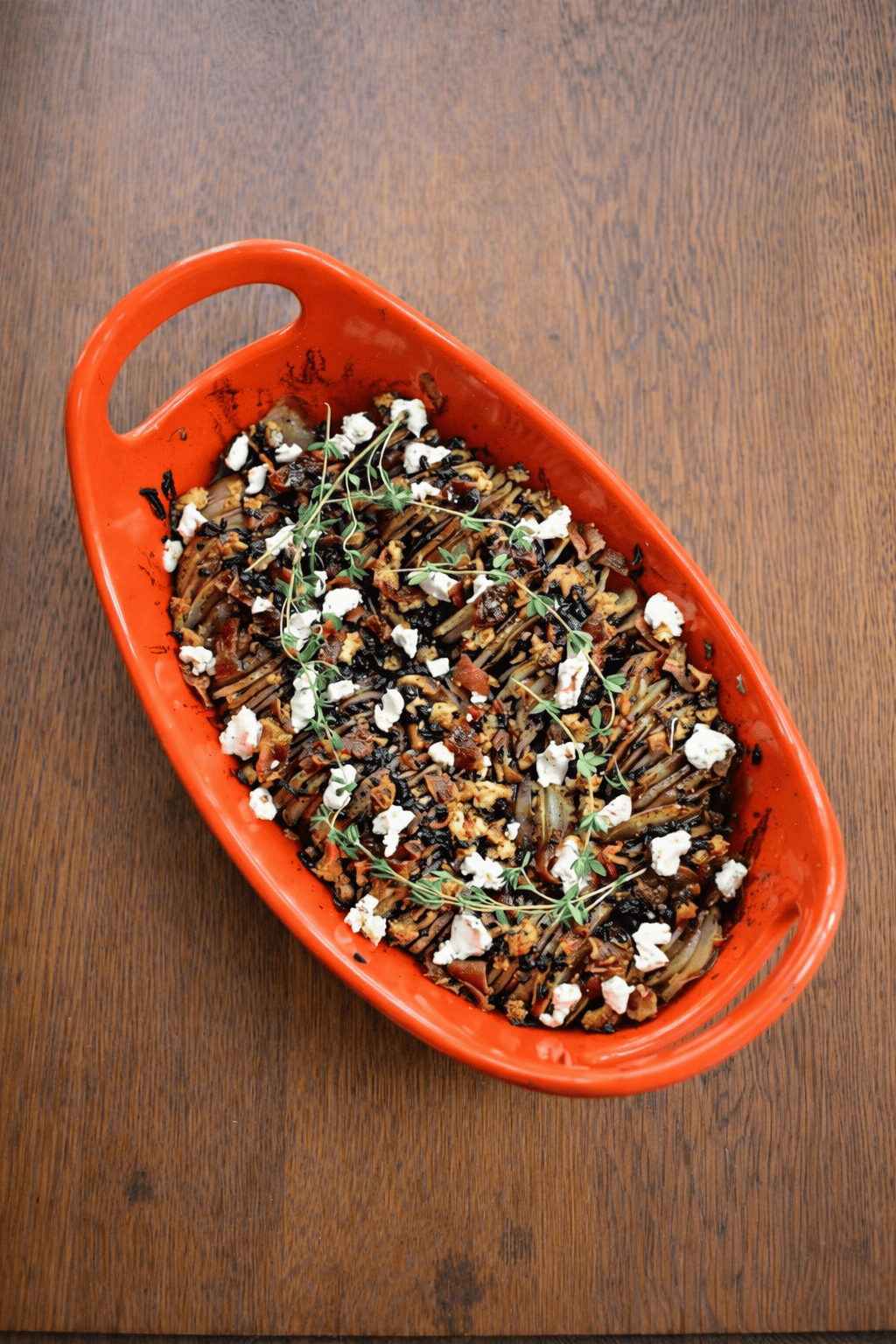 An orange baking dish with potatoes topped with bacon, goat cheese, and thyme sprigs.