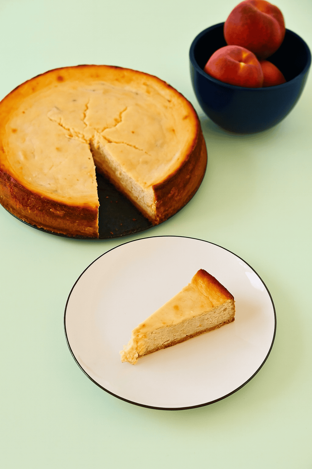 A top view of a cheesecake with a slice cut out of it, the slice of cheesecake on a plate, and a bowl of peaches.