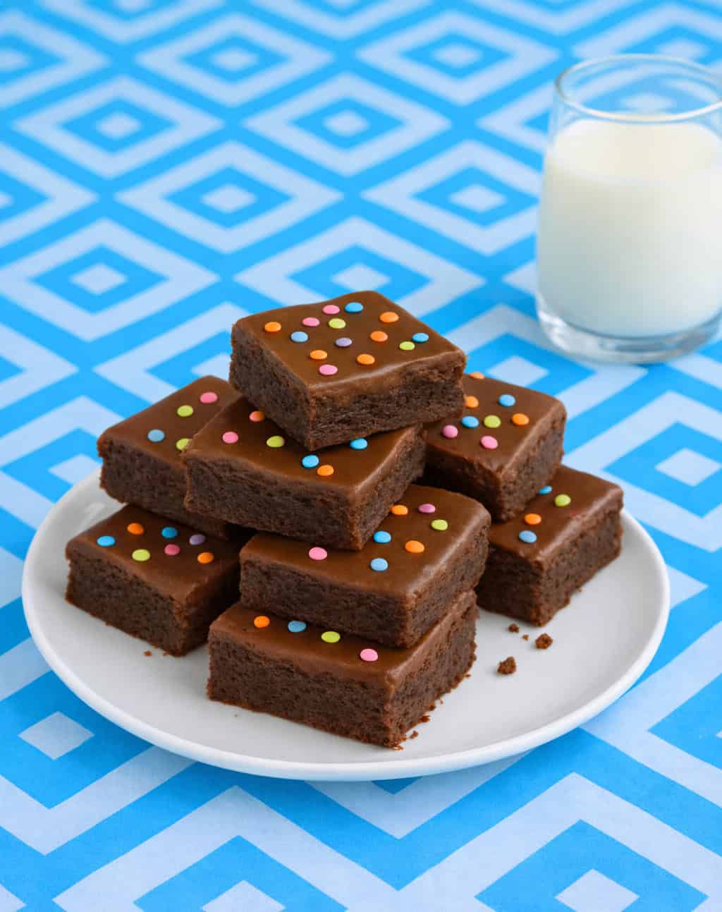 Brownies stacked on a plate with a glass of milk in the background.
