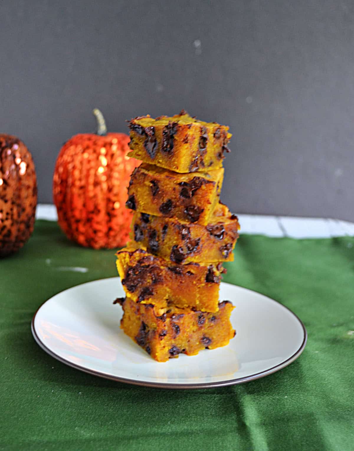 A plate with a stack of pumpkin chocolate chip bars on it and pumpkins behind the plate.