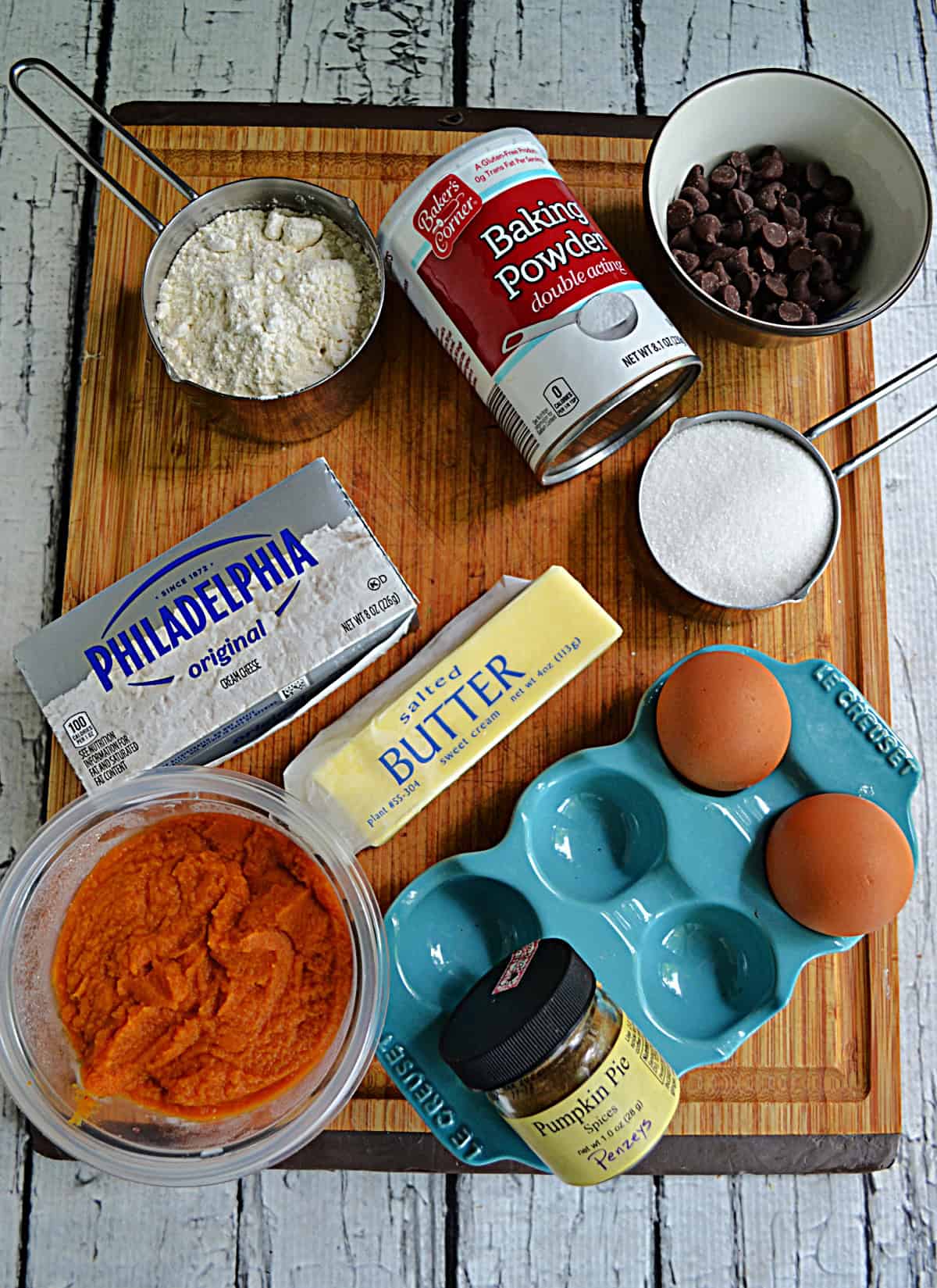 A cutting board with butter, eggs, pumpkin, cream cheese, sugar, baking powder, and chocolate chips on it.