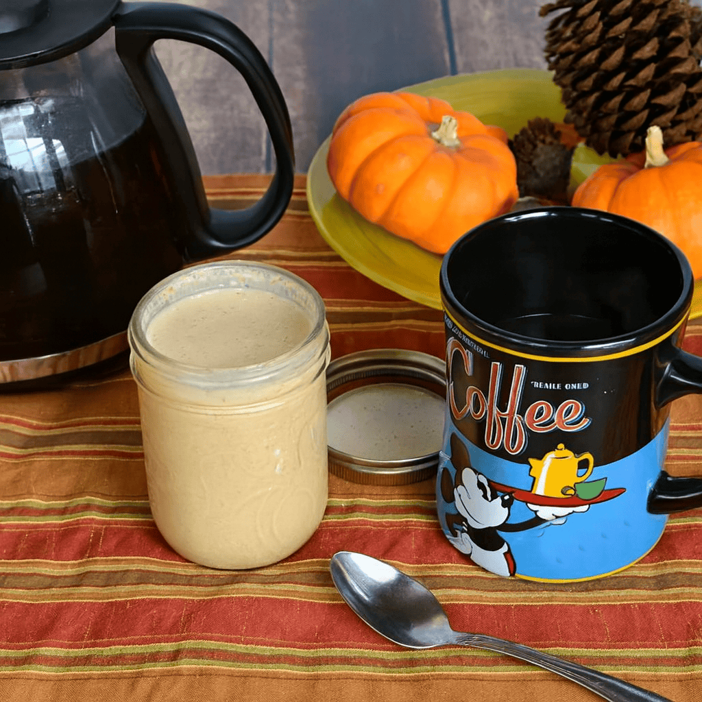 A jar of pumpkin creamer, a mug, a spoon, a coffee pot, and a plate of mini pumpkins and pinecones.