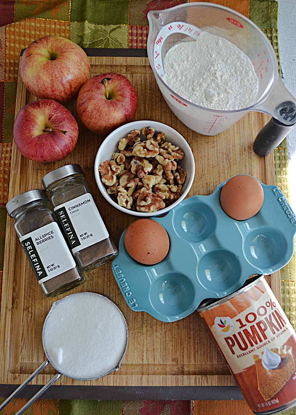 Wooden cutting board with apples, a bowl of nuts, eggs, a can of pumpkin, sugar, a cup of flour, eggs on it.