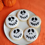 A plate of Jack Skellington cookies with pumpkins behind it.
