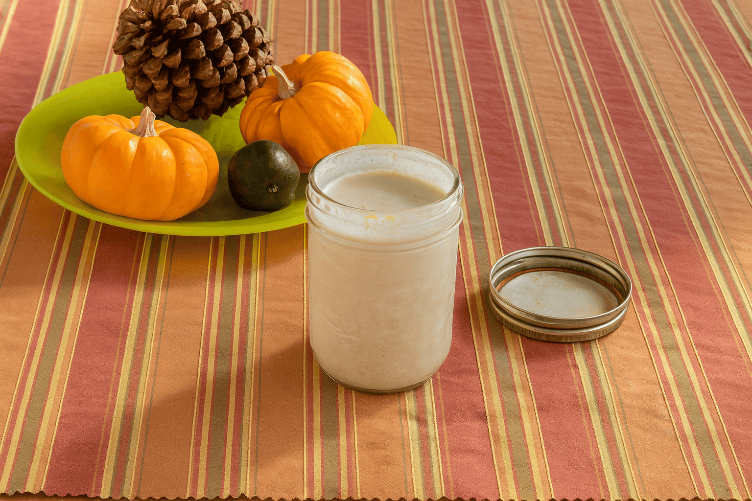 A jar of pumpkin creamer with a plate of mini pumpkins and pinecones behidn it.