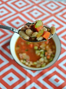 A close up of a spoon with beans and vegetables on it with a bowl of soup in the background.