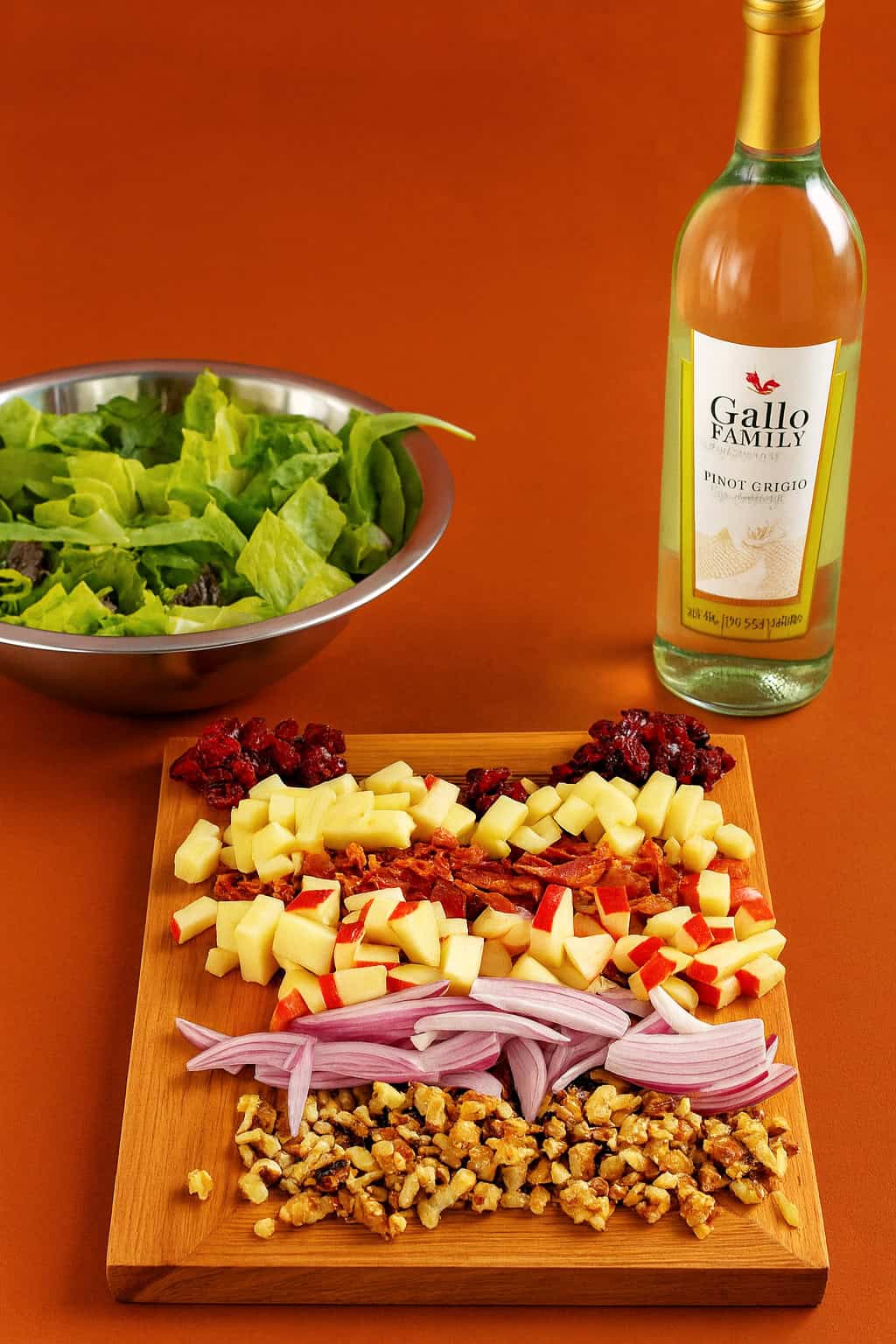 A cutting board with apples, cranberries, cheese, and onions on it with a bowl of lettuce in the background.