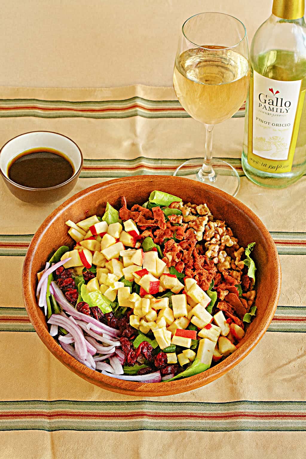 A bowl of salad topped with chopped apples, cranberries, bacon, and onions, with a bowl of dressing and a glass of wine behind it.