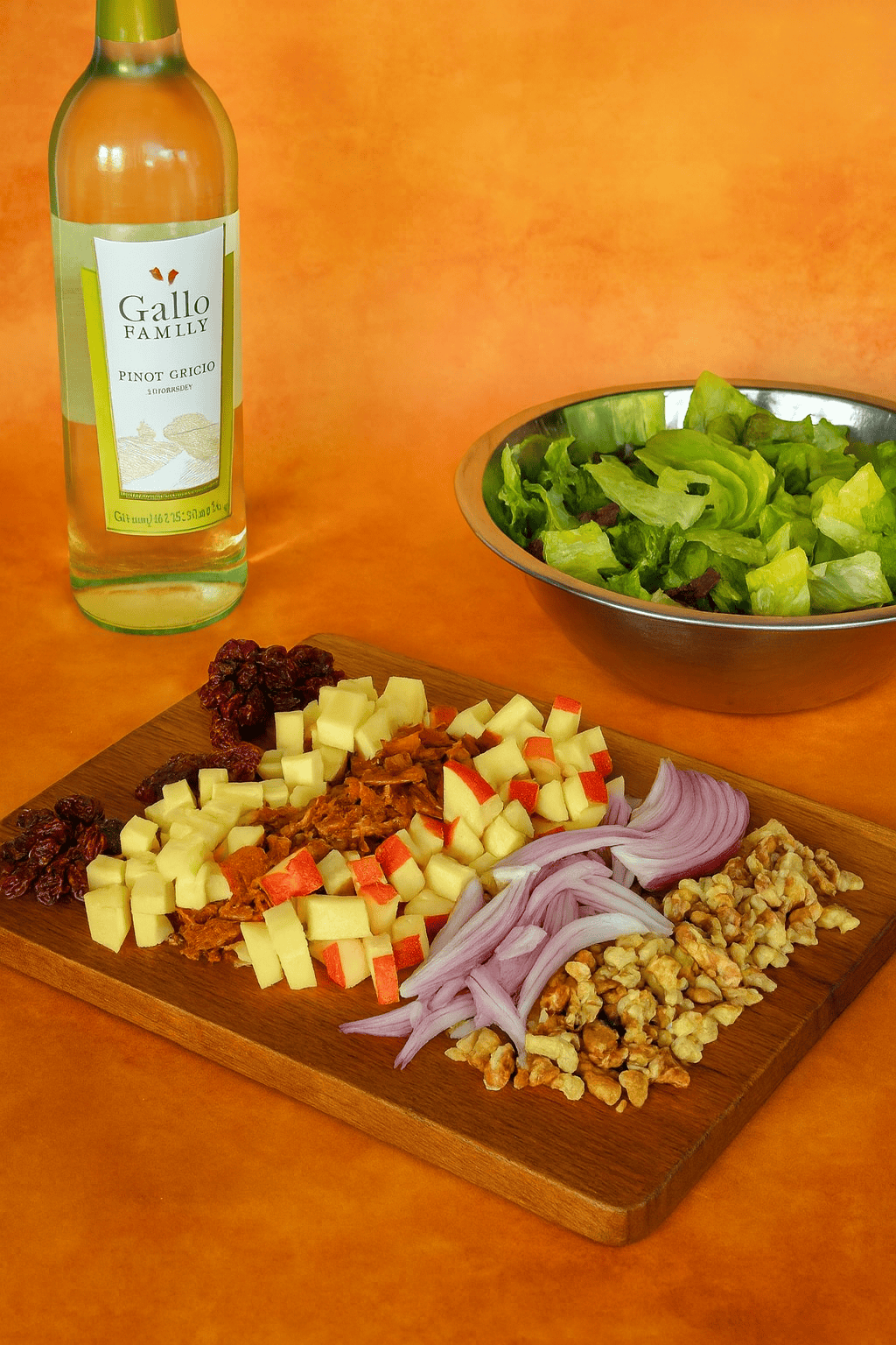 A cutting board with cranberries, apples, onions, and walnuts on it with a bowl of lettuce and a bottle of wine in the background.