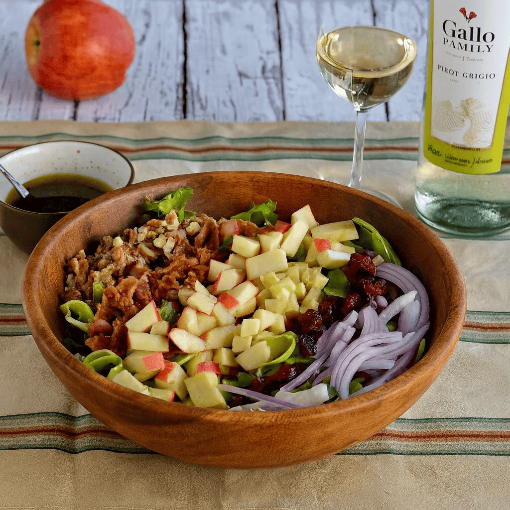 A wooden bowl of salad with chopped apples, bacon, and onions on top, an apple, a bowl of dressing, and a glass of wine behind the bowl.