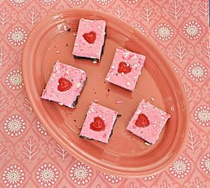 A round, pink platter with five pieces of Double Layer Double Fudge with milk chocolate on the bottom and pink white chocolate on the top each one with a heart on it on a pink background.