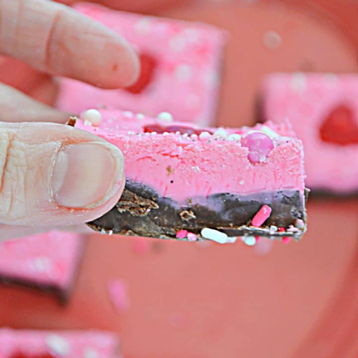 A close up of a hand holding a piece of Valentine's Day fudge with milk chocolate on the bottom and pink white chocolate on the top with a tray of fudge in the background.