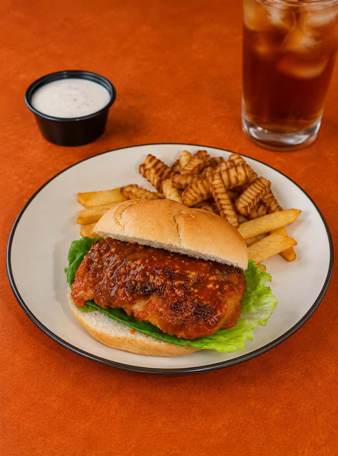 A plate with a buffalo chicken sandwich on it, a glass of iced tea and a cup of ranch dressing behind the plate.