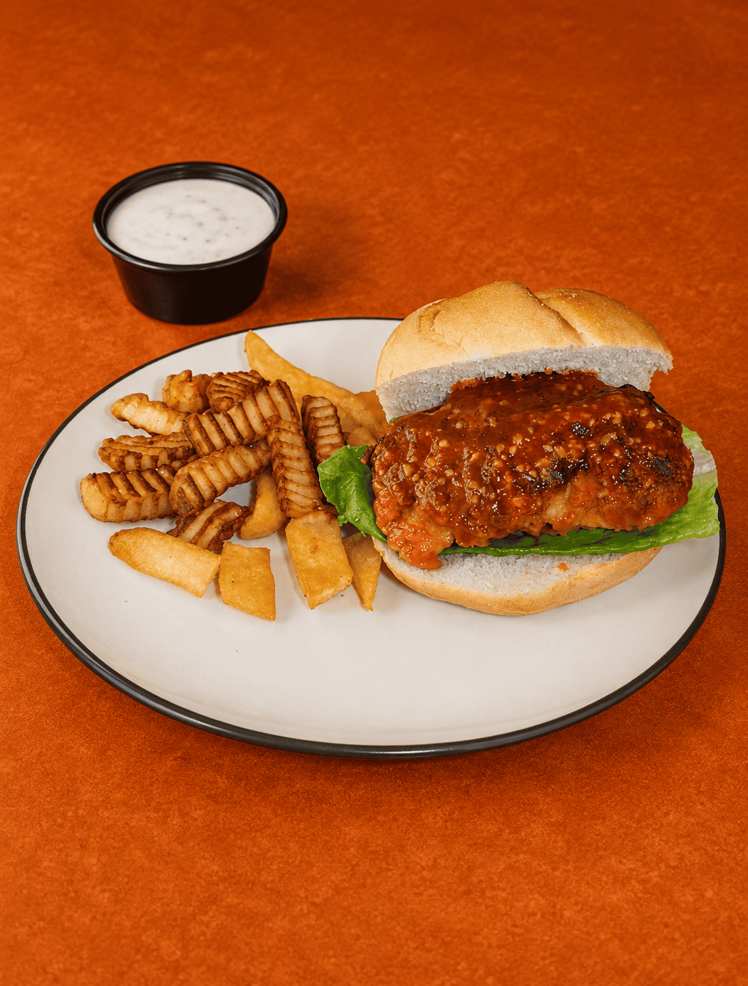 A plate with a buffalo chicken sandwich and fries on it with a cup of blue cheese behind the plate.
