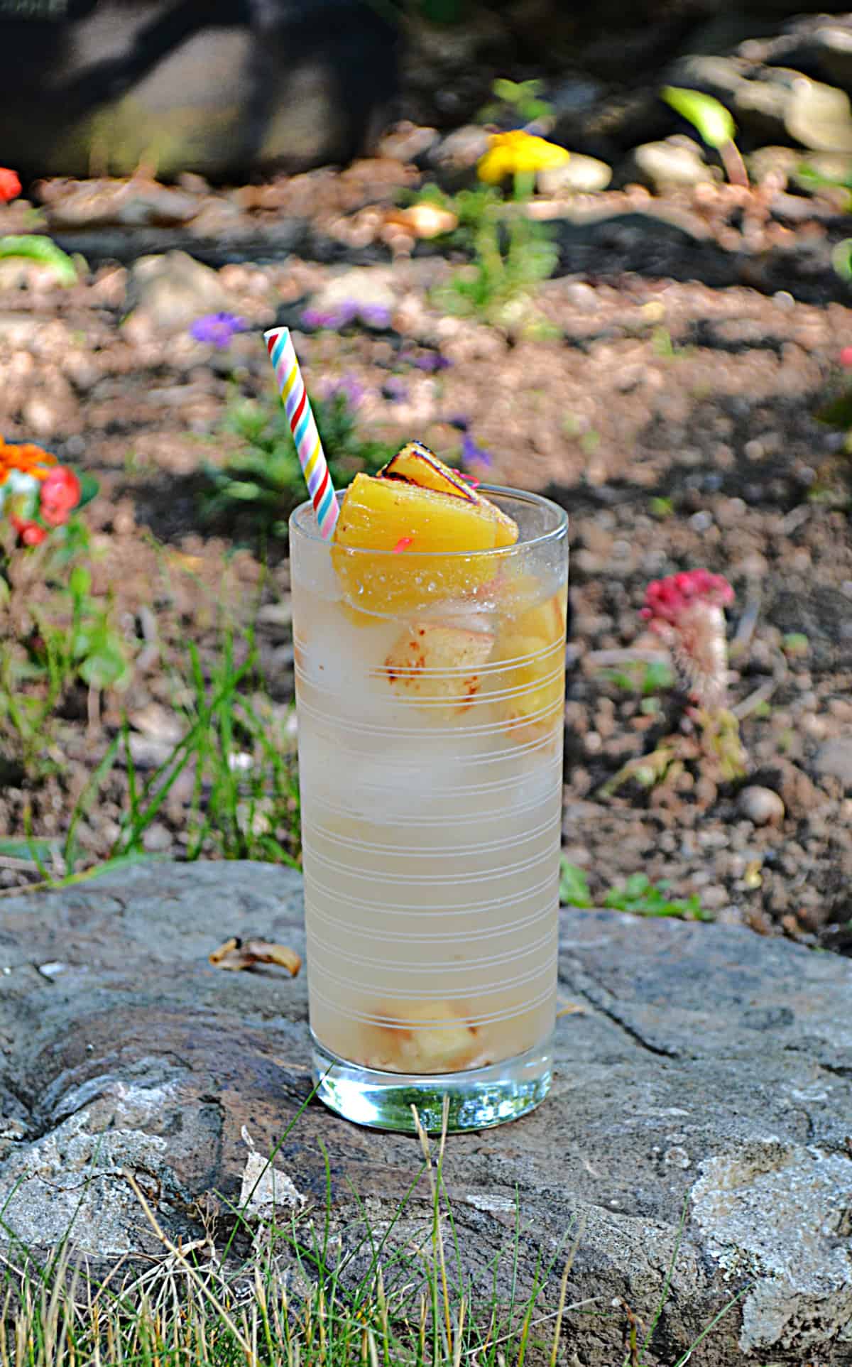 A glass of pineapple sangria on a rock in the garden with flowers behind it.
