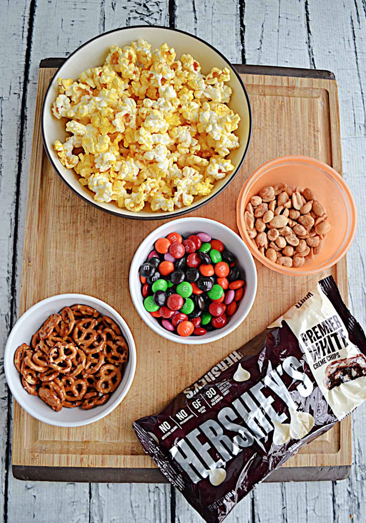 A cutting board with a bowl of popcorn, a bowl of pretzels, peanuts, M&M's, and a bag of white chocolate chips.
