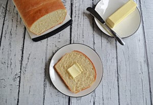 Overhead view of a plate with a piece of bread with butter on it, a loaf of bread, and a dish with butter on it.