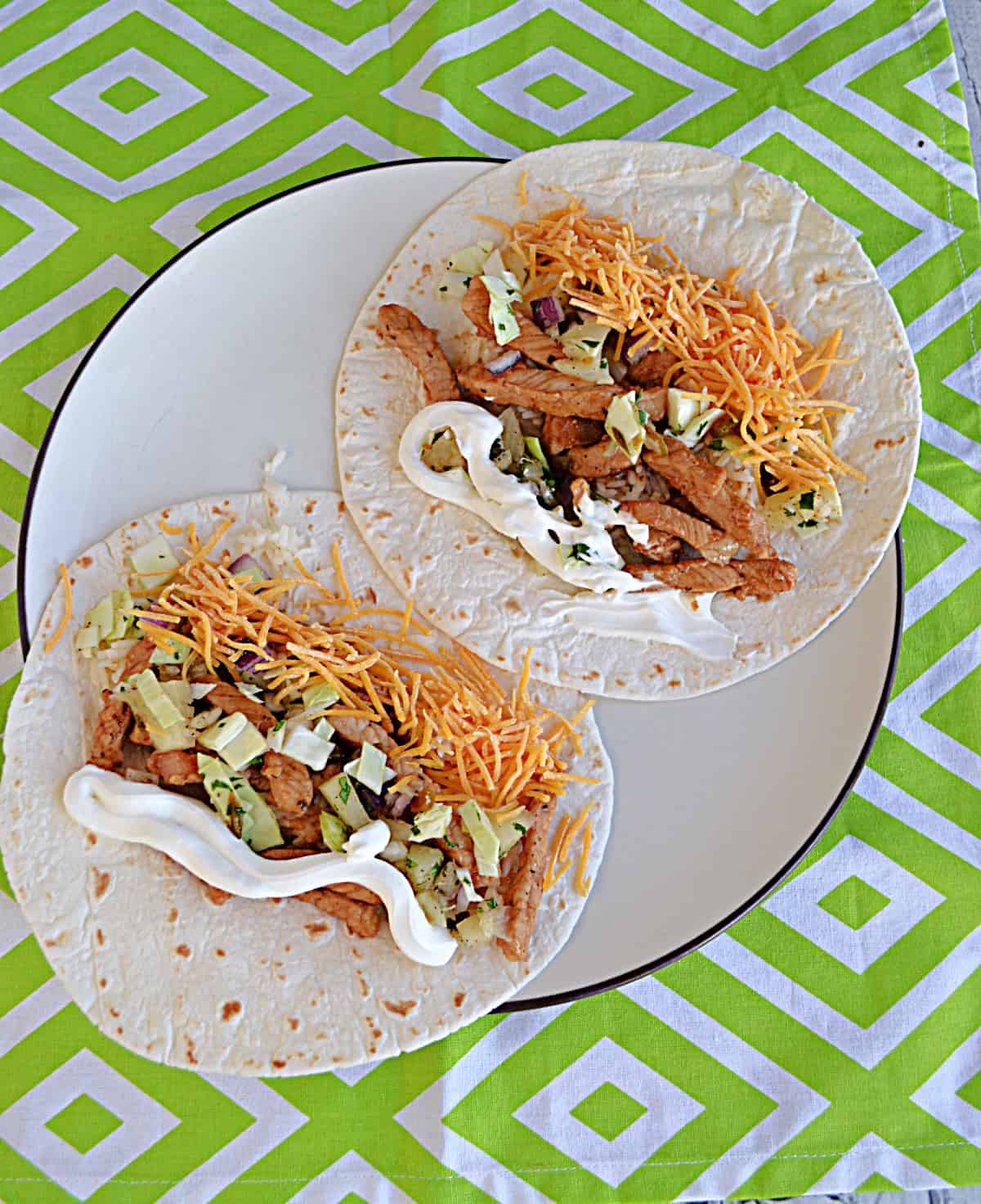 Two pork tacos topped with cheese, sour cream, and jalapeno pineapple slaw.
