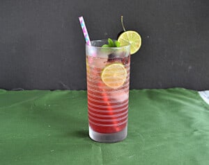 A glass of cherry mojito with a straw, lime wedge, and cherry on the side.
