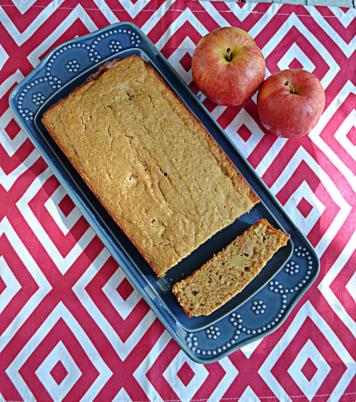 A loaf of bread on a platter with a slice cut off and two apples sitting behind the plate.