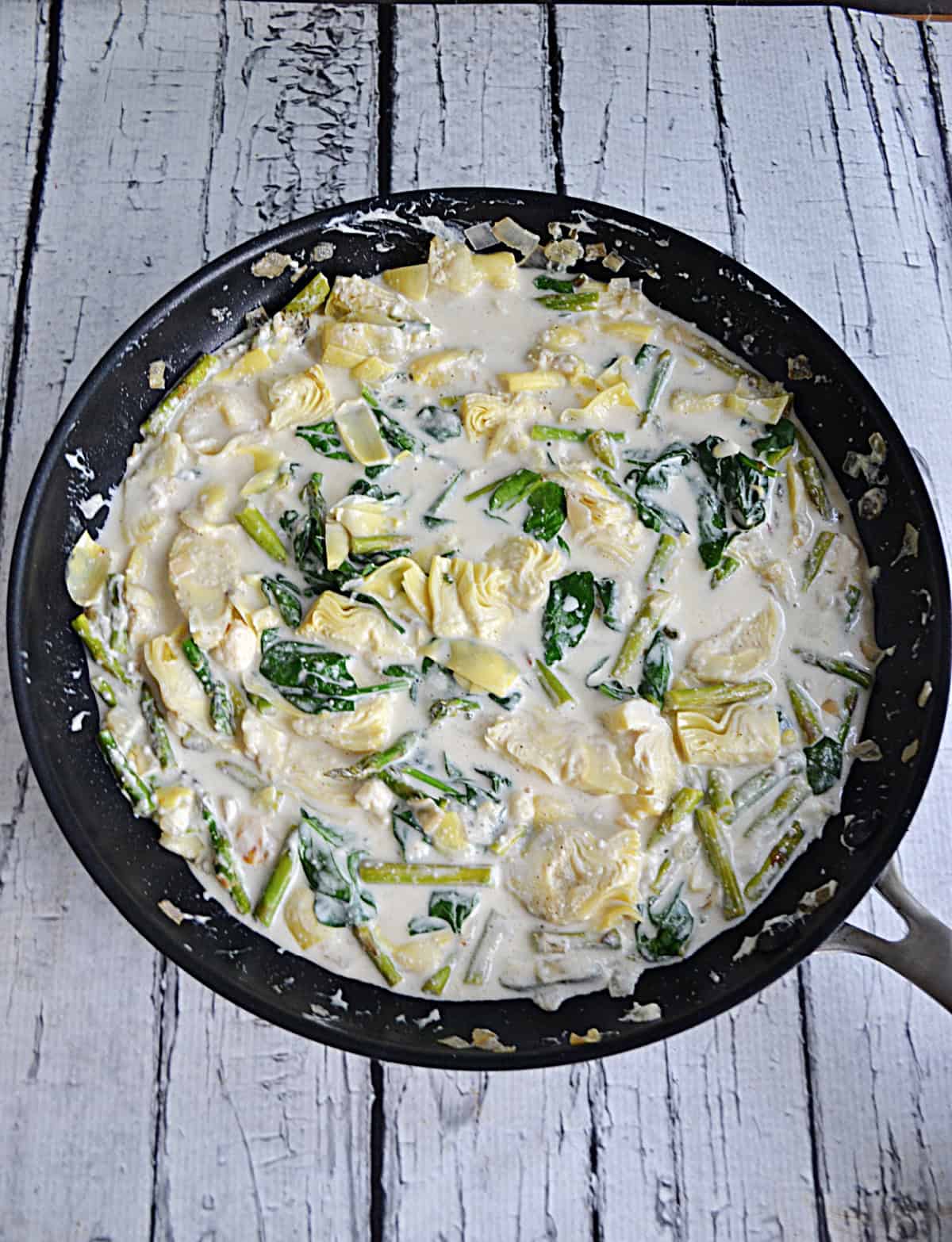 A skillet with cream sauce, spinach, artichokes, and asparagus in it.
