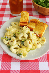 A plate of tortellini and garlic bread with a salad bowl behind the plate and a glass of iced tea.