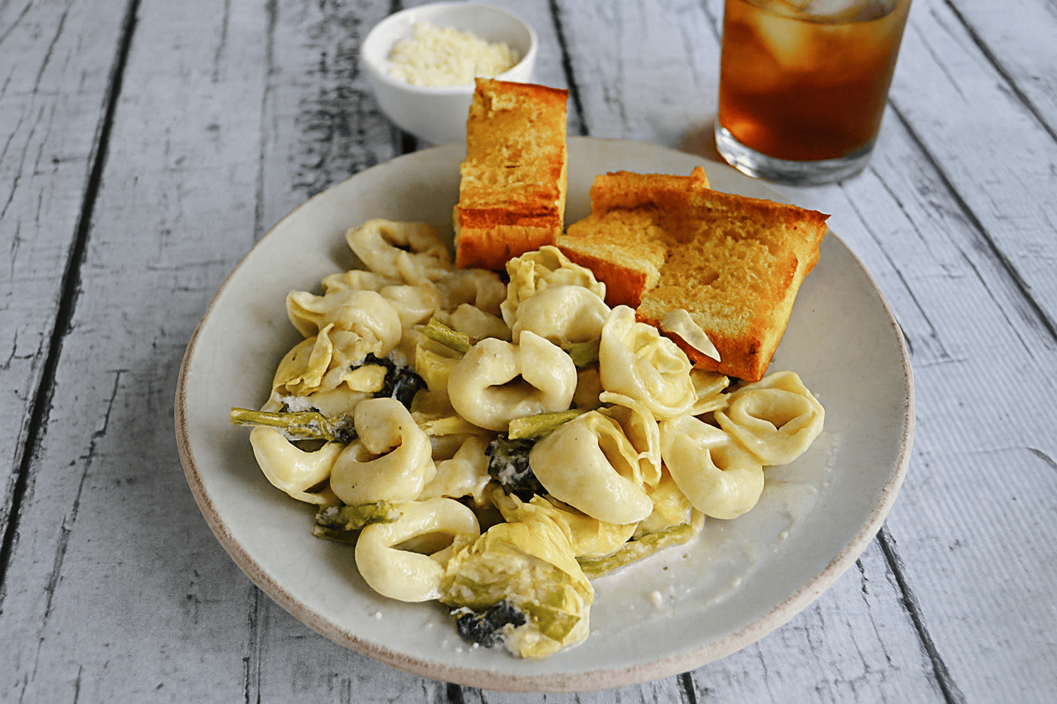 A plate of tortellini with slices of garlic bread, a bowl of parmesan cheese, and a glass of iced tea.