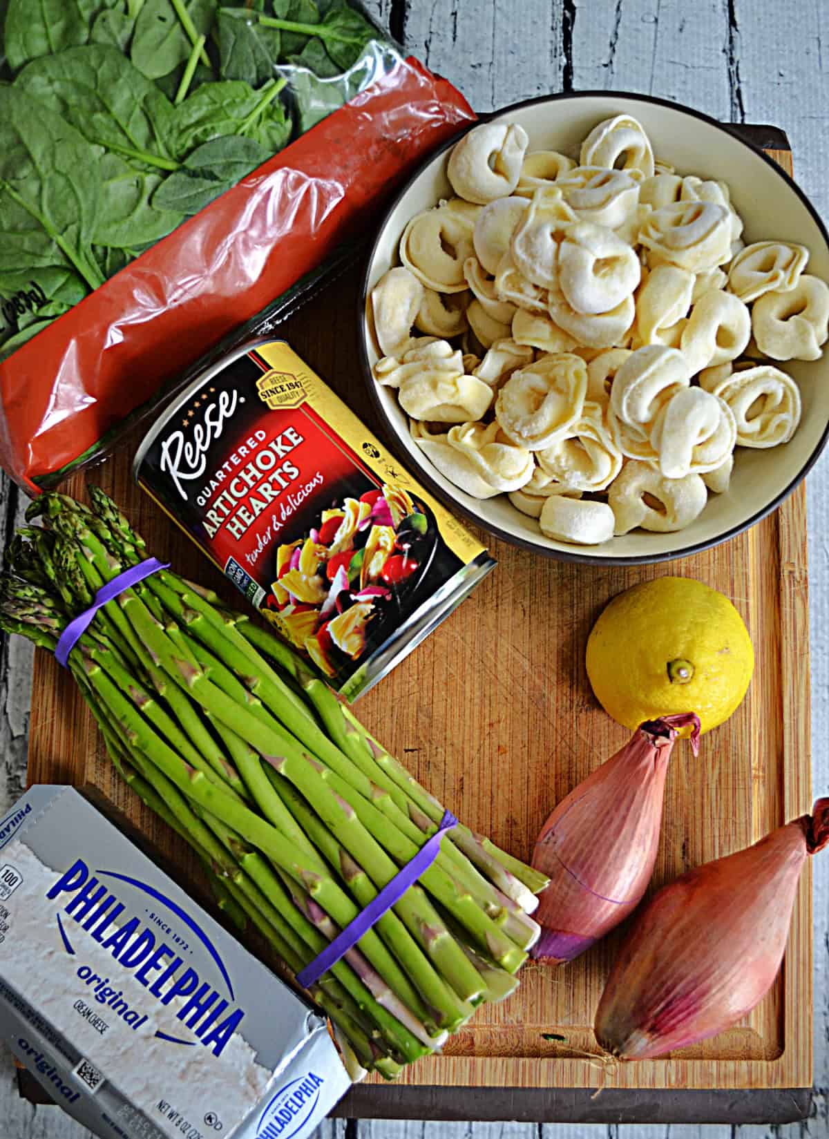 A cutting board with tortellini, asparagus, cream cheese, artichokes, spinach, and lemon on it.