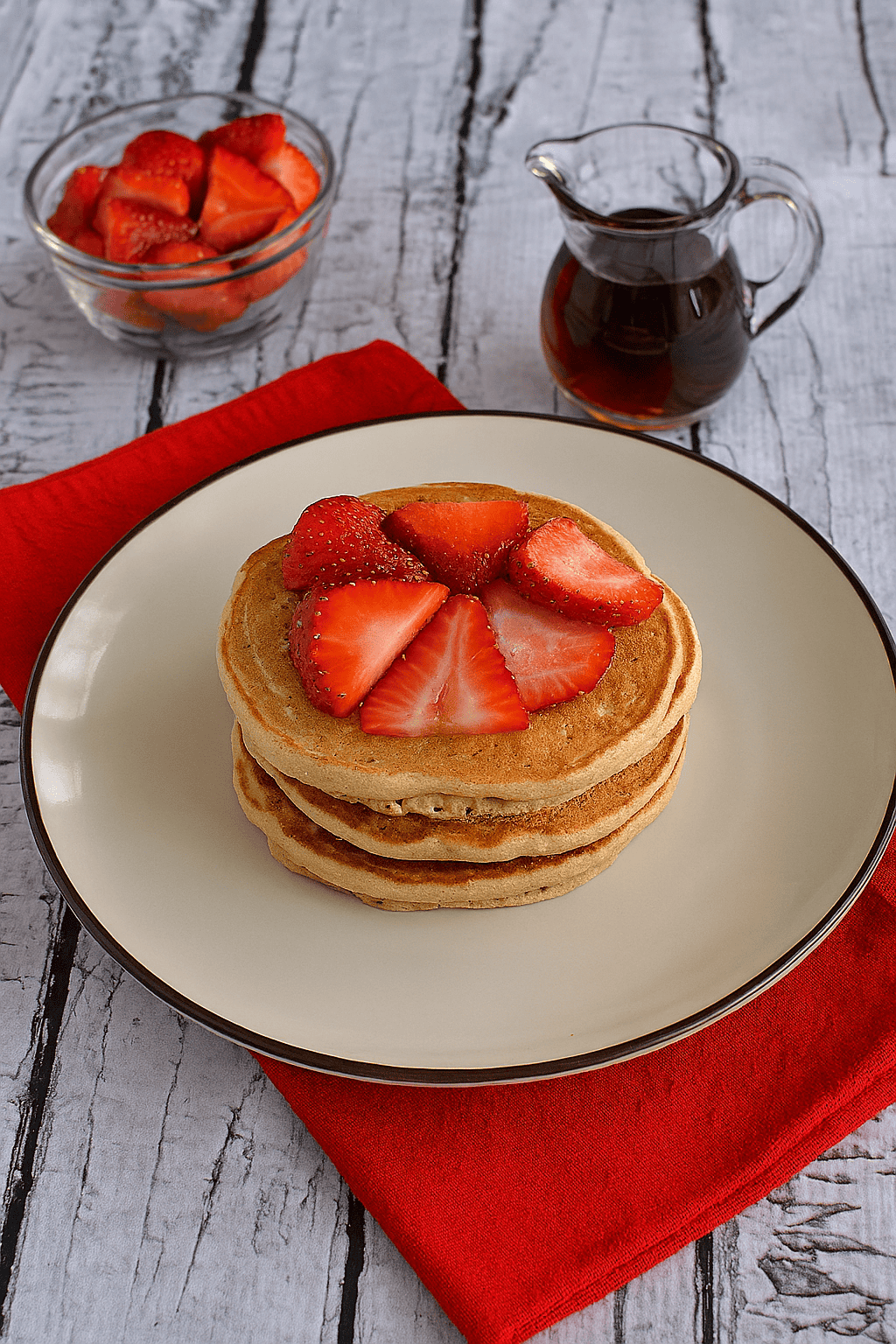 A plate of pancakes with sliced strawberries on top and a dish of strawberries in the background.