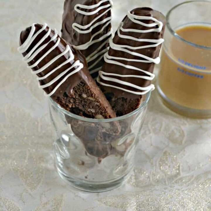 A glass with 3 biscotti in it that are dipped in chocolate and a mug of coffee behind them.