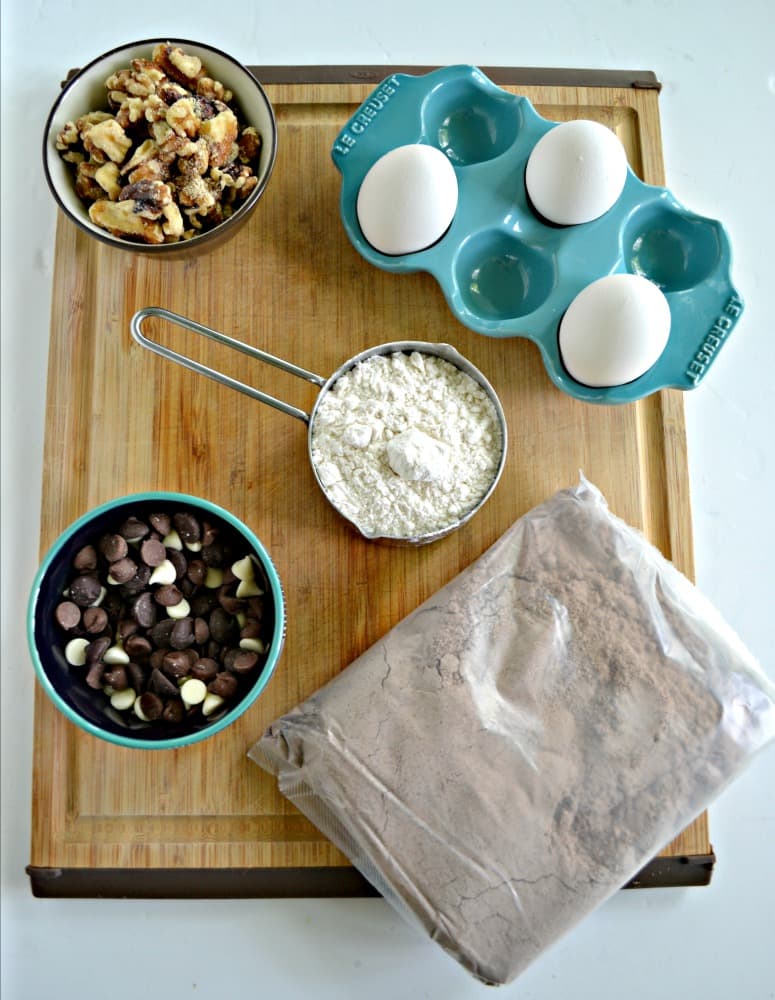 A cutting board with a brownie mix, a cup of flour, a bowl of chocolate chips, a bowl of walnuts, and 3 eggs on it.