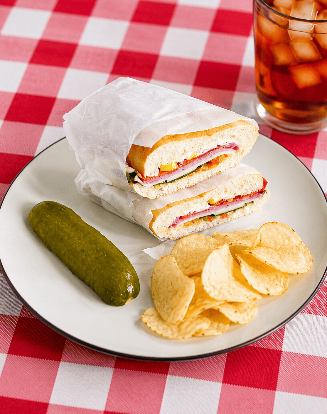 A plate with two wrapped Muffuletta sandwich halves, a pickle, and a handful of chips.